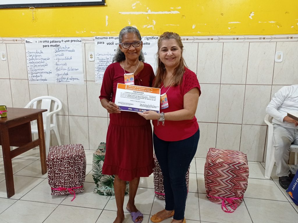 Aposentada Maria Lucia recebendo certificado das mãos da oficineira Rita Figueiredo